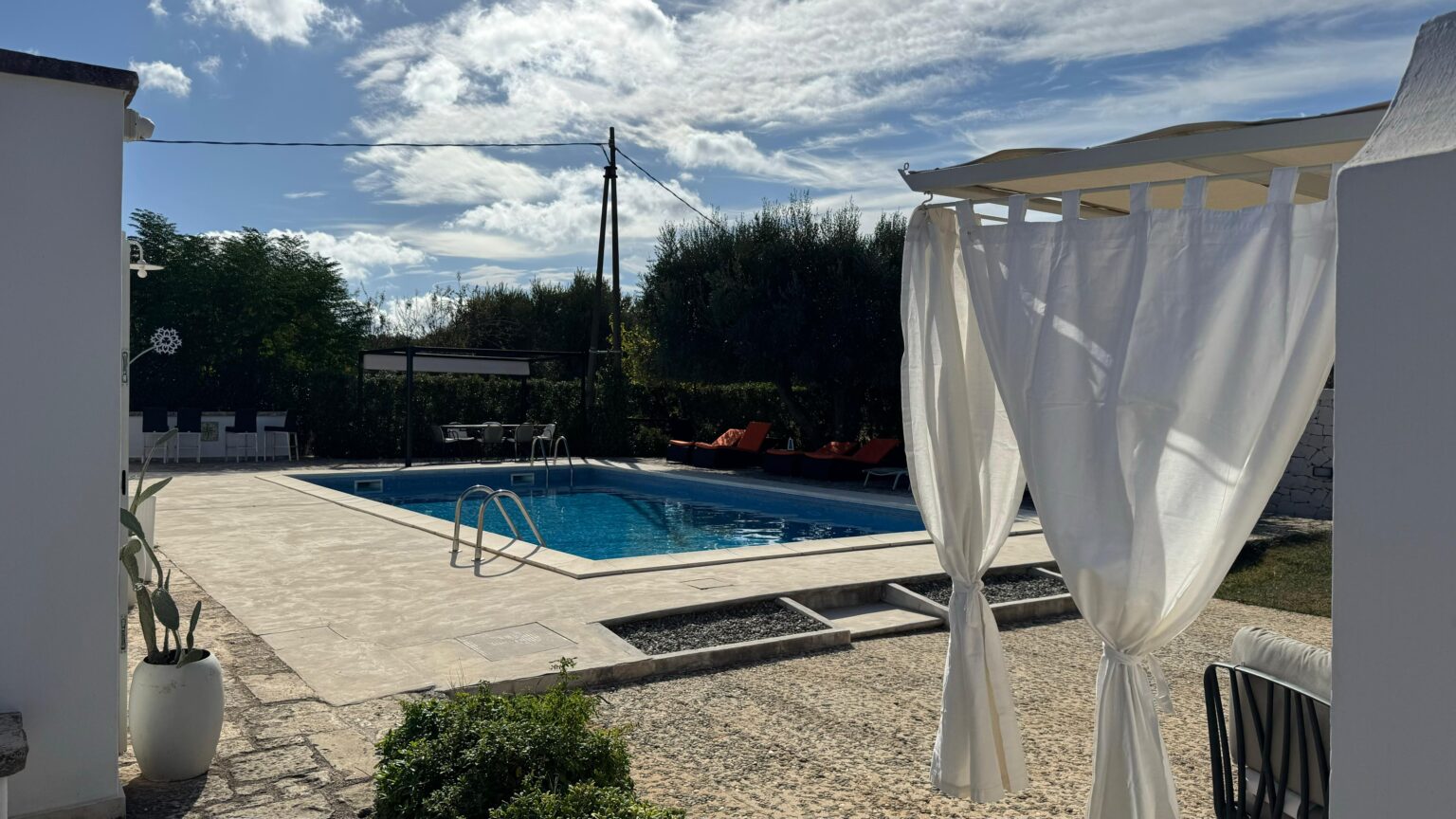 Outdoor swimming pool surrounded by a landscaped area, featuring a white draped cabana and lounge chairs, under a clear blue sky with scattered clouds. | Only on SopranoVillas