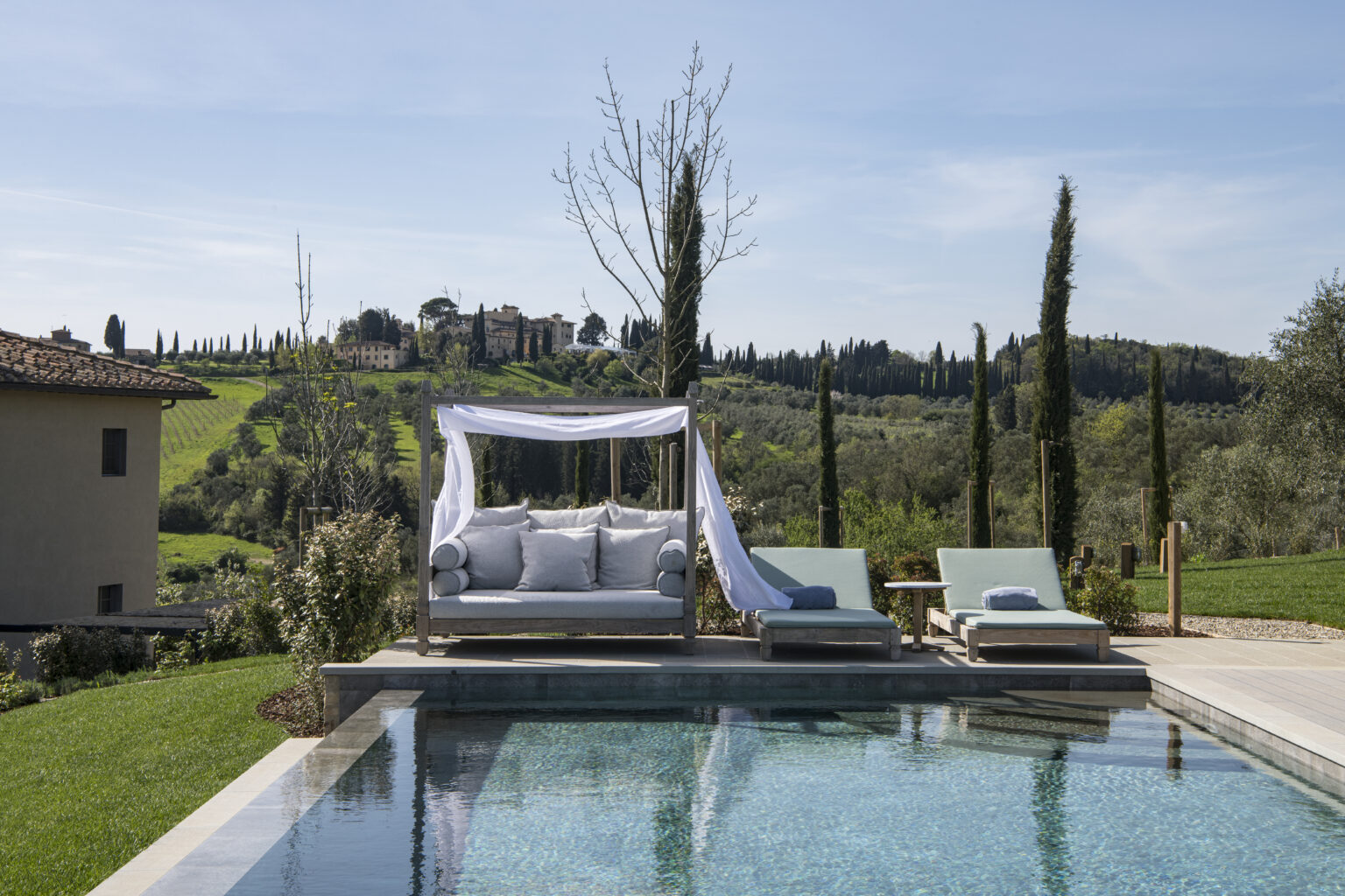 Luxurious outdoor lounge area by a serene pool, featuring a shaded daybed and sun loungers, with lush green hills and cypress trees in the background, perfect for relaxation and leisure in a scenic landscape. | Only on SopranoVillas