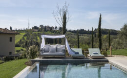 Luxurious outdoor lounge area by a serene pool, featuring a shaded daybed and sun loungers, with lush green hills and cypress trees in the background, perfect for relaxation and leisure in a scenic landscape.
