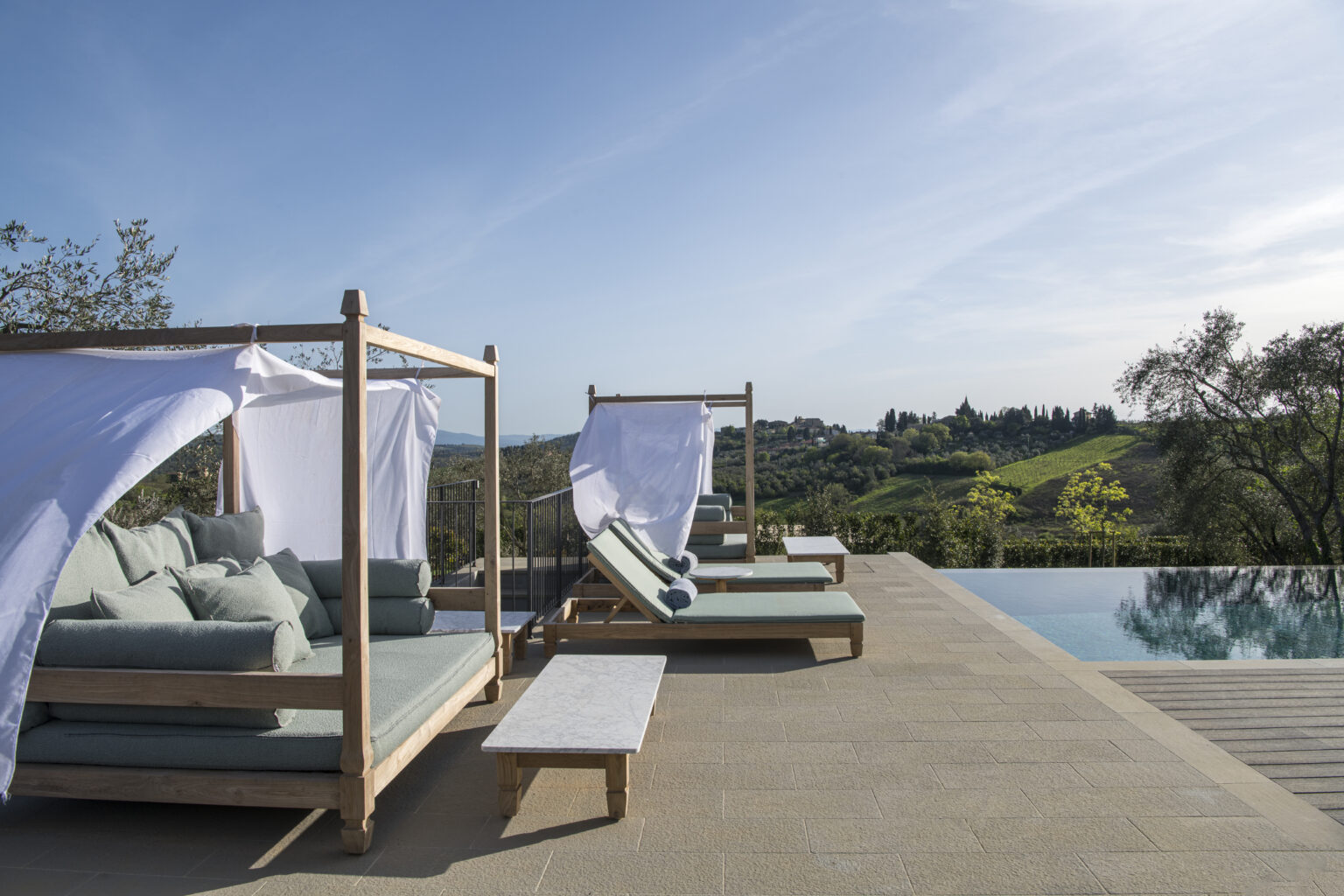 Luxurious outdoor lounge area with shaded daybeds beside a sparkling swimming pool, overlooking lush green hills and a clear blue sky. Ideal for relaxation and enjoying scenic views. | Only on SopranoVillas