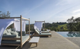 Luxurious outdoor lounge area with shaded daybeds beside a sparkling swimming pool, overlooking lush green hills and a clear blue sky. Ideal for relaxation and enjoying scenic views.