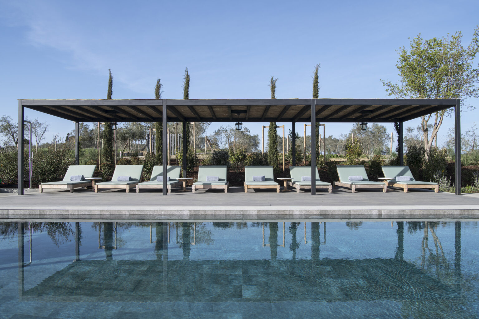Modern outdoor pool area featuring a shaded pergola and comfortable lounge chairs surrounded by greenery and blue skies. Ideal for relaxation and leisure. | Only on SopranoVillas