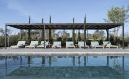 Modern outdoor pool area featuring a shaded pergola and comfortable lounge chairs surrounded by greenery and blue skies. Ideal for relaxation and leisure.