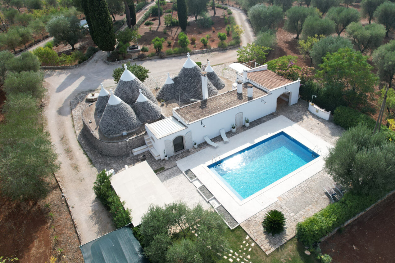 Aerial view of a picturesque villa featuring traditional trulli architecture, surrounded by olive trees and a swimming pool, set in a serene landscape. | Only on SopranoVillas