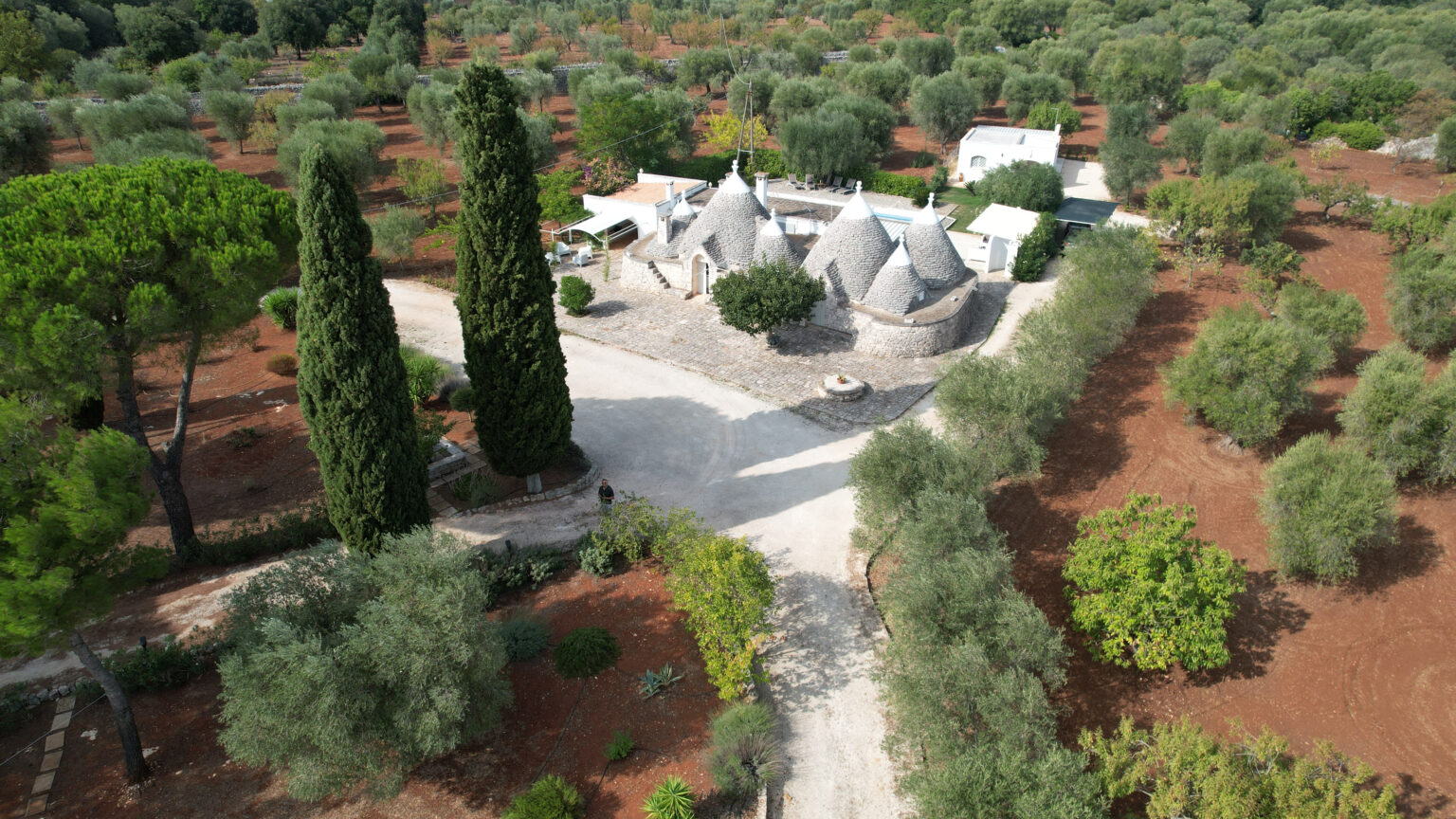Aerial view of a traditional trullo house surrounded by lush olive trees and landscaped gardens, showcasing a serene rural setting in Italy. | Only on SopranoVillas