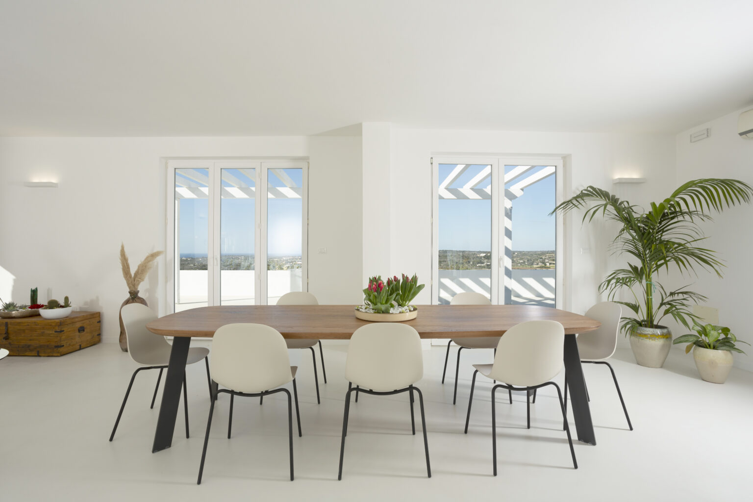 Modern dining room featuring a large wooden table surrounded by white chairs, with large windows offering a view of the outdoors. The space is illuminated by natural light and decorated with indoor plants, creating a bright and airy atmosphere. | Only on SopranoVillas