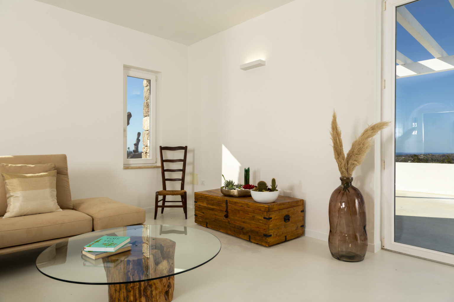 Modern living room interior featuring a beige sofa, a glass coffee table, a wooden storage chest, and a decorative vase with dried plants. Large window provides natural light and a view of the outdoors. | Only on SopranoVillas
