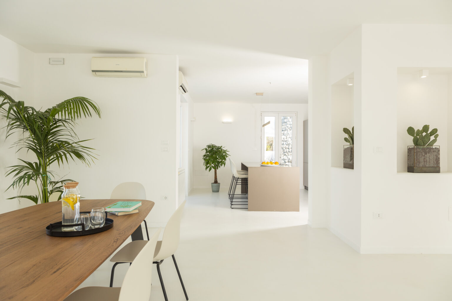Modern open-concept living space featuring a wooden dining table with glassware and books, a stylish kitchen island, and indoor plants, all set against a bright, minimalist white backdrop. | Only on SopranoVillas