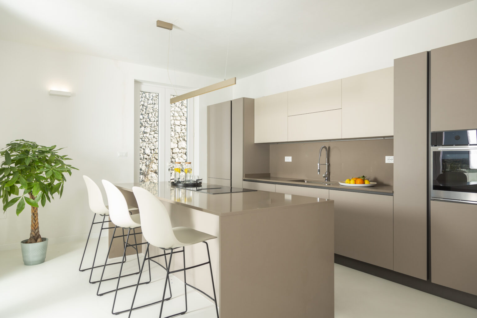 Modern kitchen design featuring minimalist cabinetry in neutral tones, a spacious island with bar stools, and a potted plant, providing a bright and inviting atmosphere. | Only on SopranoVillas