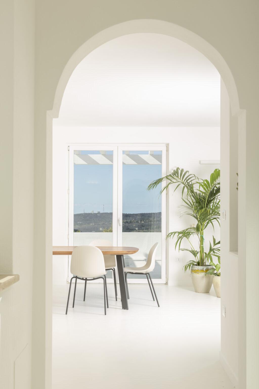 Bright and modern dining area featuring a wooden table and minimalist chairs, framed by an archway. Large windows offer a view of the outdoors, with a green potted plant adding a touch of nature to the interior. | Only on SopranoVillas