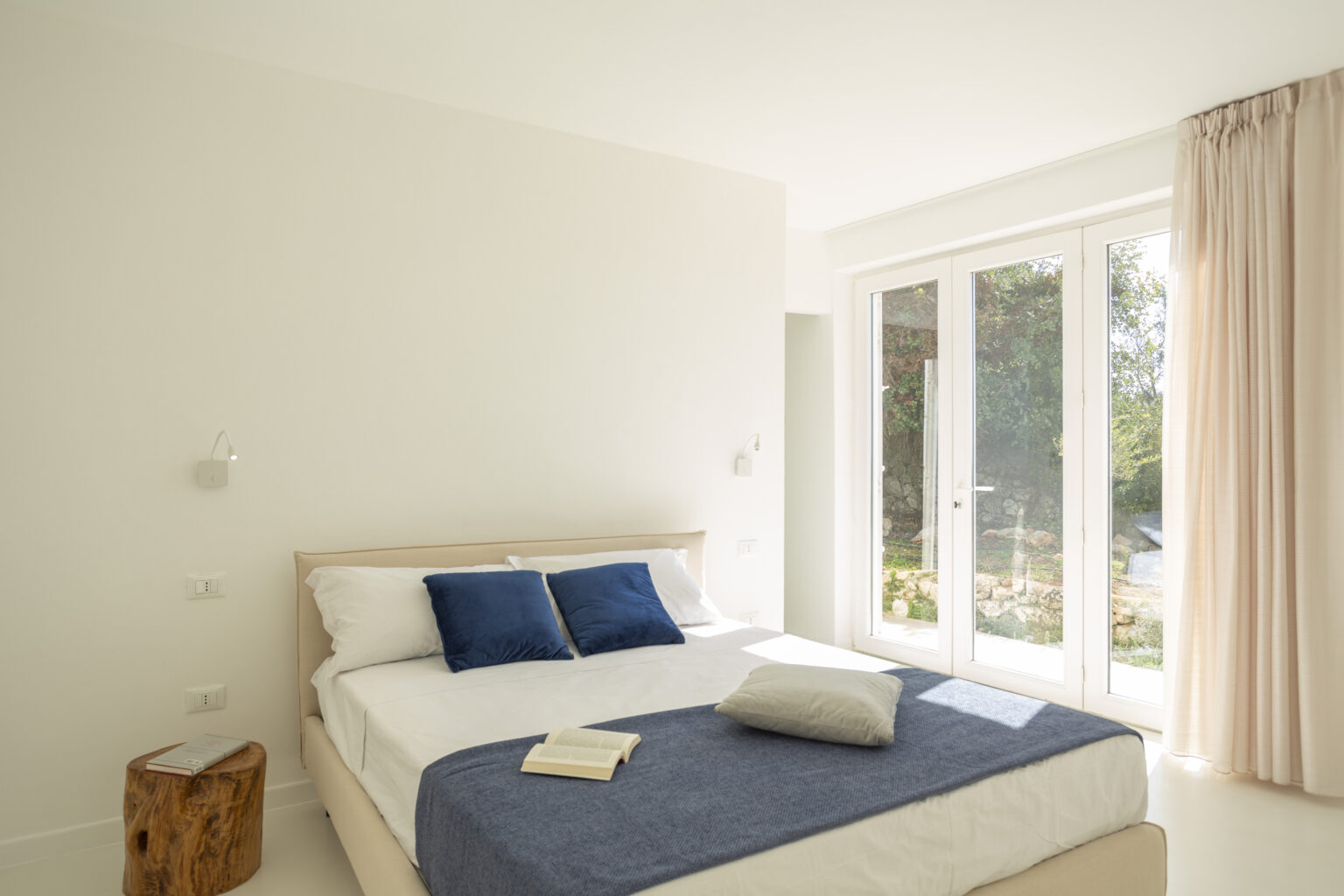 Modern bedroom featuring a queen-sized bed with blue accent pillows, a cozy throw blanket, and a wooden side table. Bright natural light floods the room through large glass doors that lead to an outdoor garden space. Minimalist design with white walls and soft curtains creates a serene atmosphere. | Only on SopranoVillas