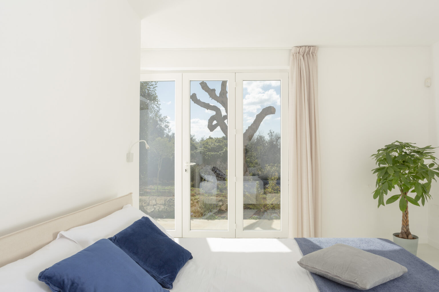Bright and airy bedroom featuring a large window with a view of a garden. The room includes a bed with blue pillows, a soft gray throw pillow, and a potted plant, creating a serene and inviting atmosphere. | Only on SopranoVillas