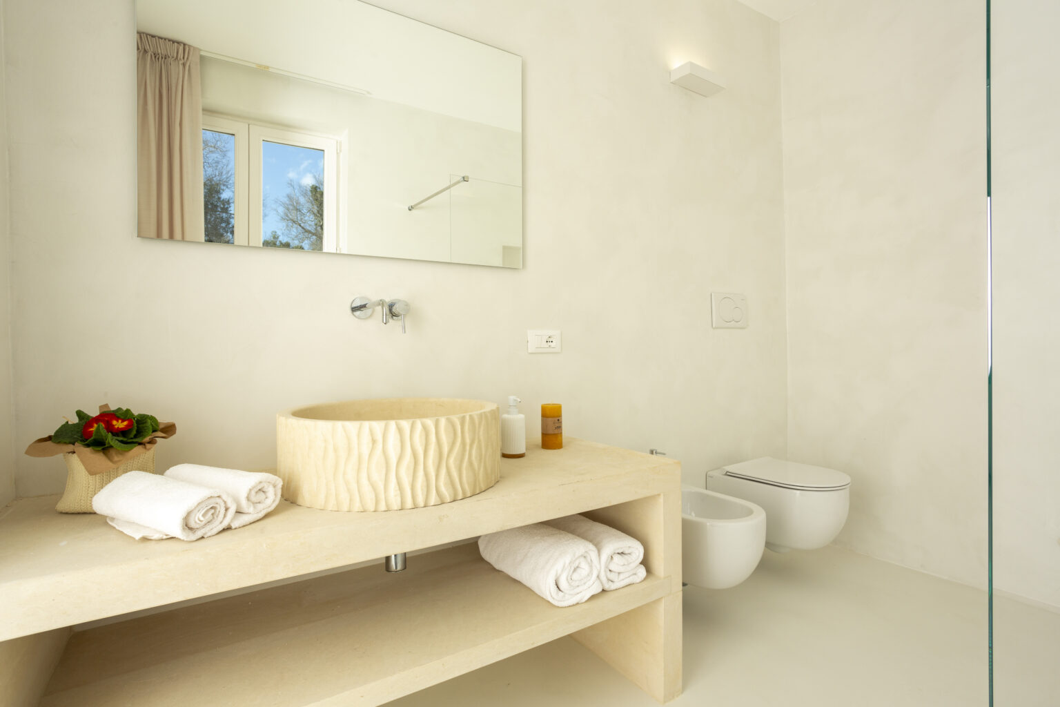 Modern bathroom interior featuring a minimalist design with a stone sink, neatly rolled white towels, and decorative plants. The space includes a large mirror, natural light from a window, and contemporary fixtures, creating a serene and stylish atmosphere. | Only on SopranoVillas