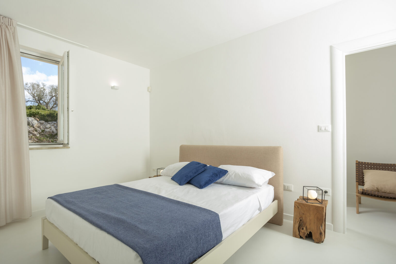 Modern bedroom featuring a queen-size bed with white linens and blue accents, minimalistic decor, natural light from a window, and a cozy seating area. Ideal for relaxation and comfort. | Only on SopranoVillas