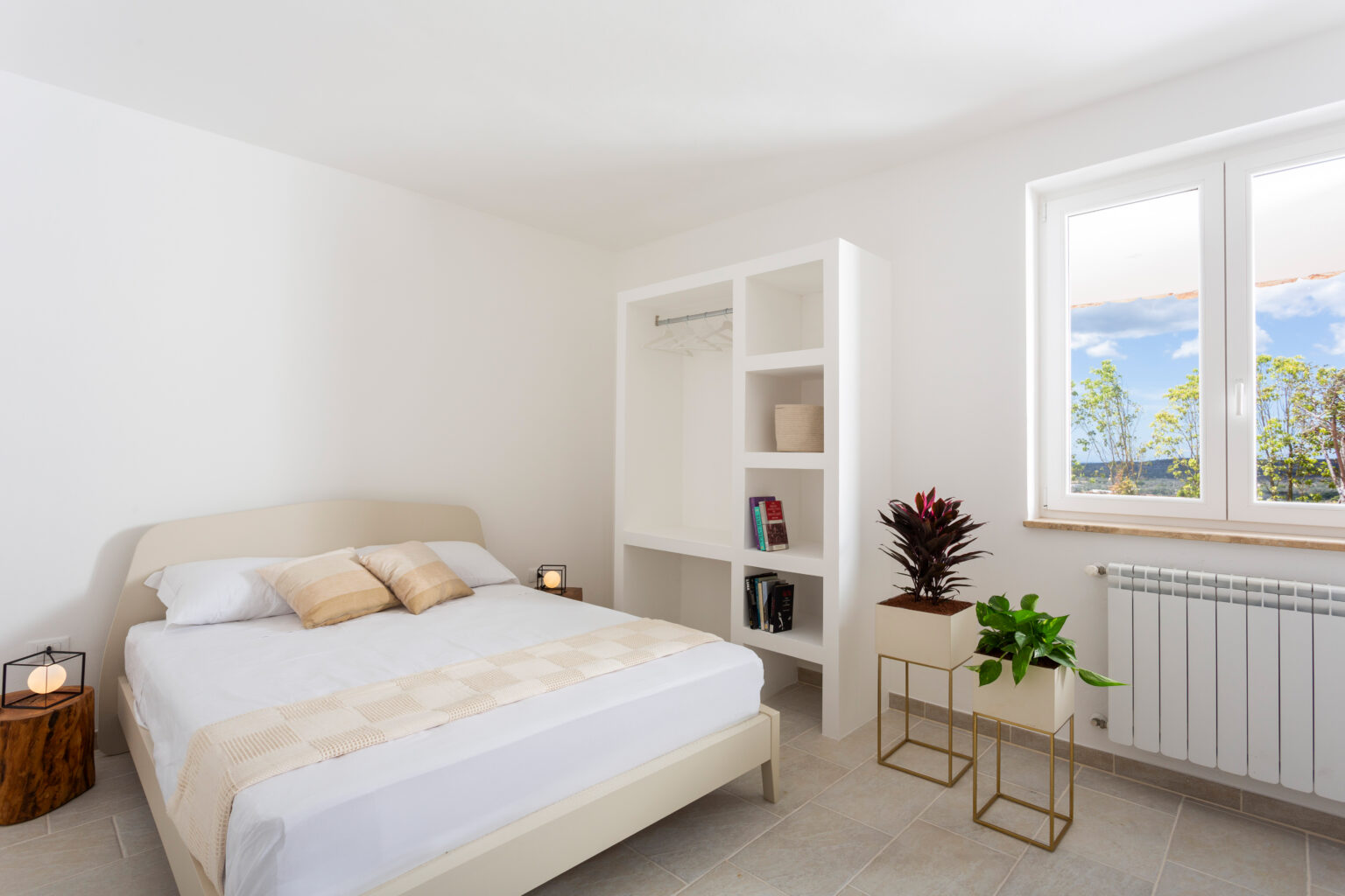 Bright and modern bedroom featuring a comfortable bed with neutral bedding, a minimalist bookshelf, and a stylish plant accent. Large windows allow natural light to fill the space, enhancing the clean, airy atmosphere. Ideal for home decor inspiration. | Only on SopranoVillas