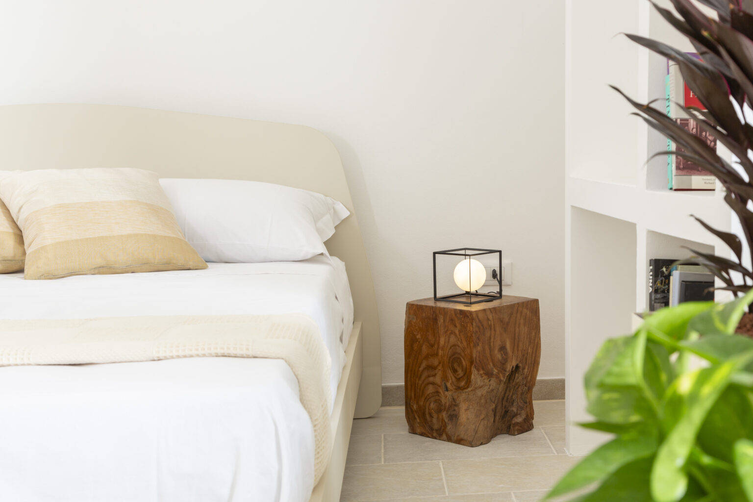 Cozy bedroom featuring a light-colored bed with decorative pillows, a rustic wooden nightstand with a lantern, and a green plant in the foreground, creating a serene and inviting atmosphere. | Only on SopranoVillas
