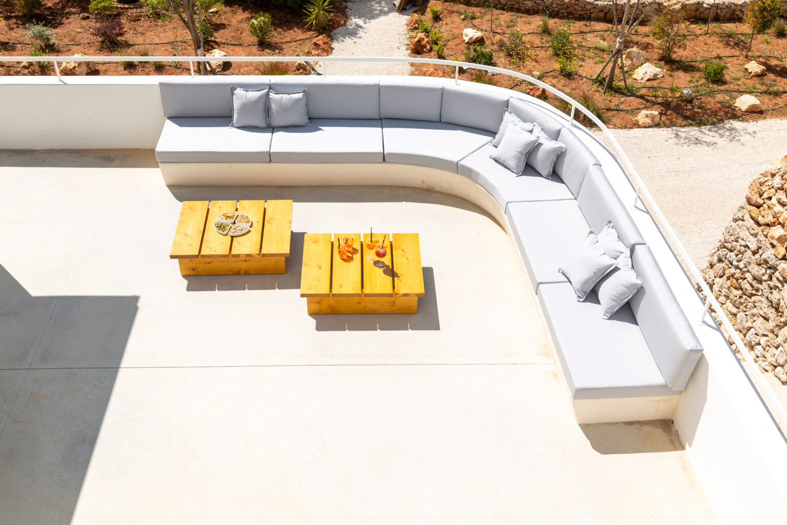 Modern outdoor lounge area featuring a curved gray sectional sofa with decorative pillows and two wooden coffee tables on a concrete patio, surrounded by landscaped greenery. | Only on SopranoVillas