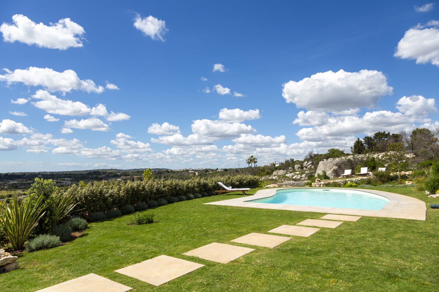 Scenic outdoor pool area with lush green grass, stone pathway, and vibrant blue sky dotted with fluffy clouds, set against a backdrop of rolling hills and greenery. Ideal for relaxation and leisure activities. | Only on SopranoVillas