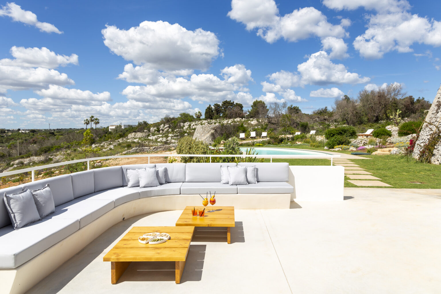 Modern outdoor lounge area featuring a curved sectional sofa and wooden coffee tables, surrounded by lush greenery and a swimming pool under a bright blue sky with fluffy clouds. Perfect for relaxation and entertaining in a serene landscape. | Only on SopranoVillas