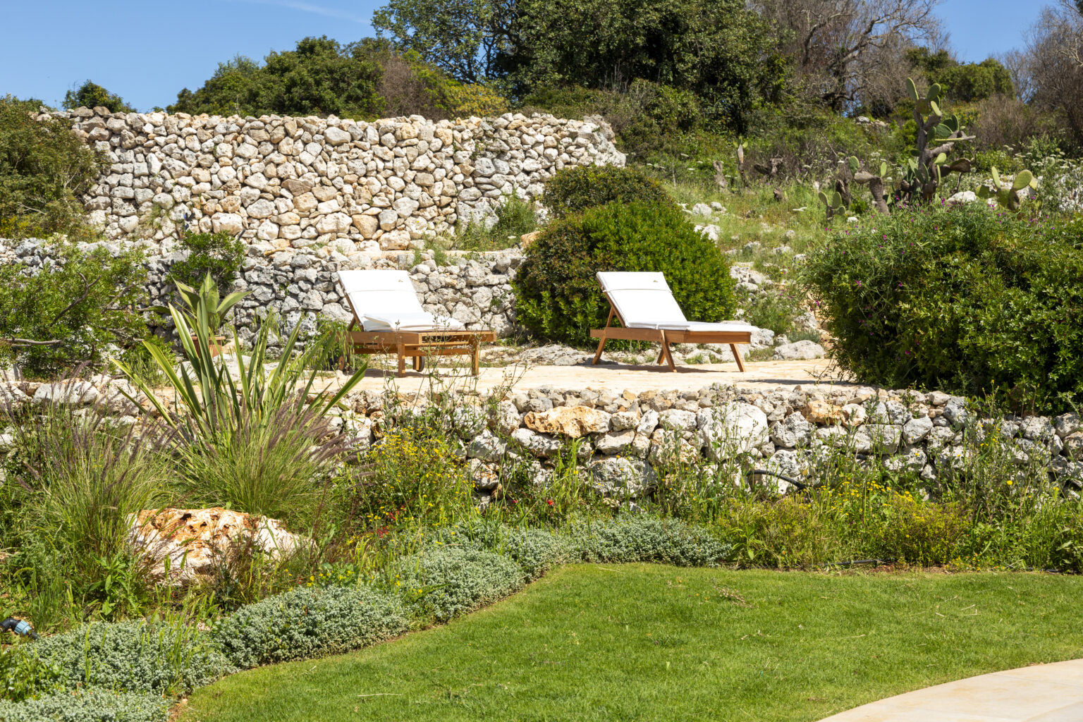 Sun loungers on a stone terrace surrounded by lush greenery and colorful flowers in a serene garden setting. | Only on SopranoVillas