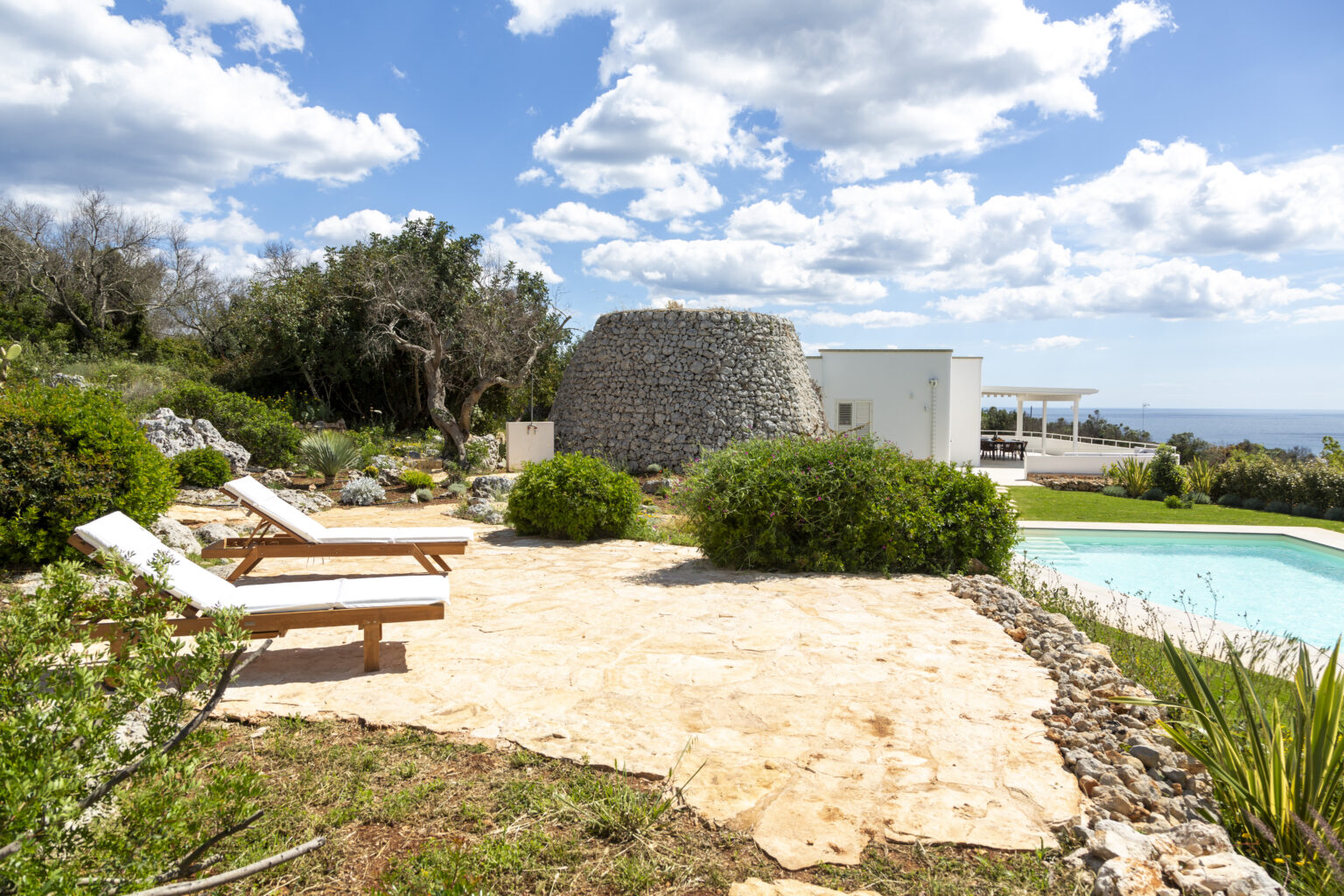 Luxurious outdoor pool area featuring sun loungers, lush greenery, and a unique stone structure, set against a bright blue sky with scattered clouds. Ideal for relaxation and enjoying scenic views. | Only on SopranoVillas