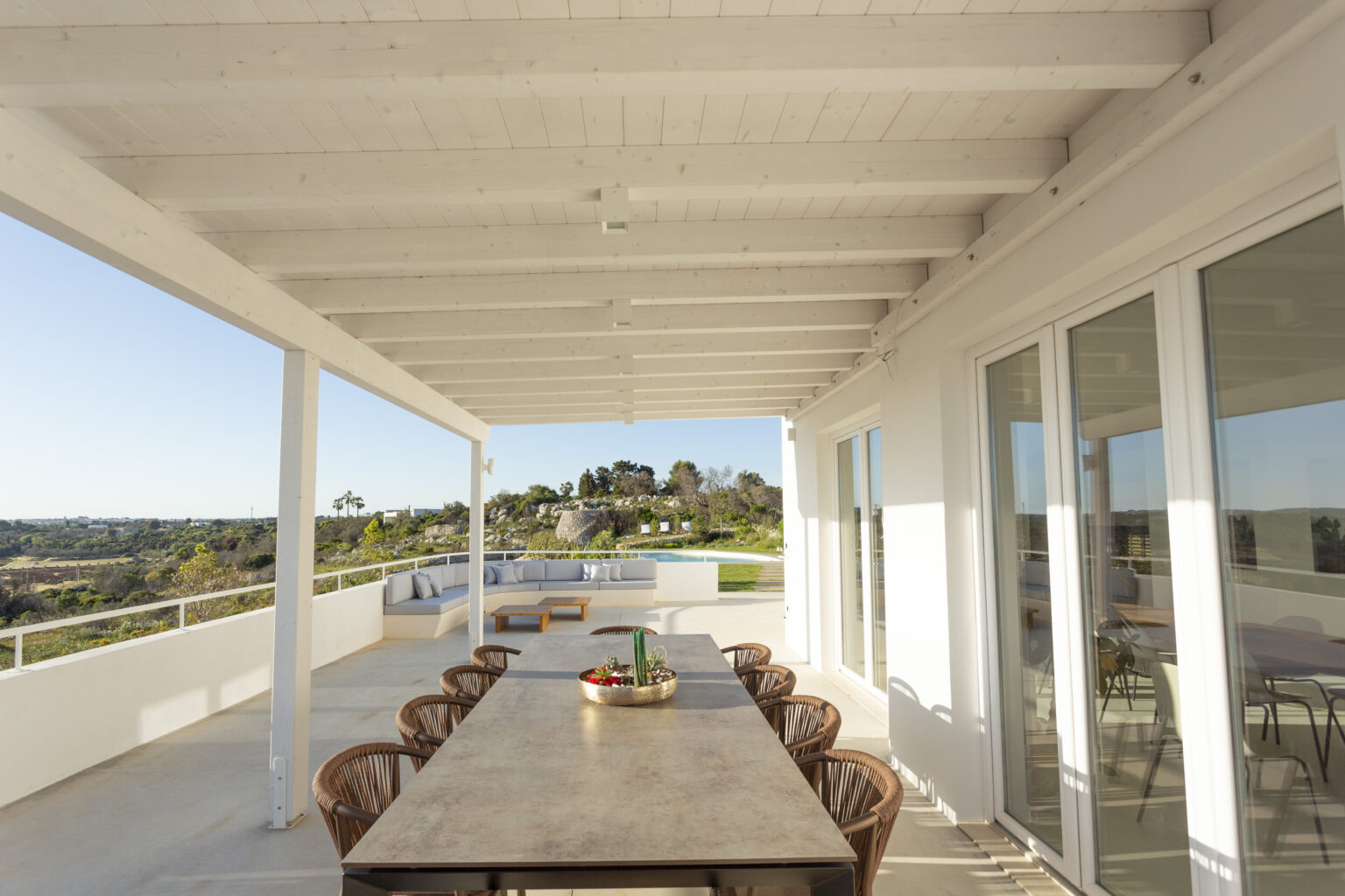 Spacious outdoor dining area with a long wooden table and wicker chairs, overlooking a scenic landscape under a bright blue sky. | Only on SopranoVillas
