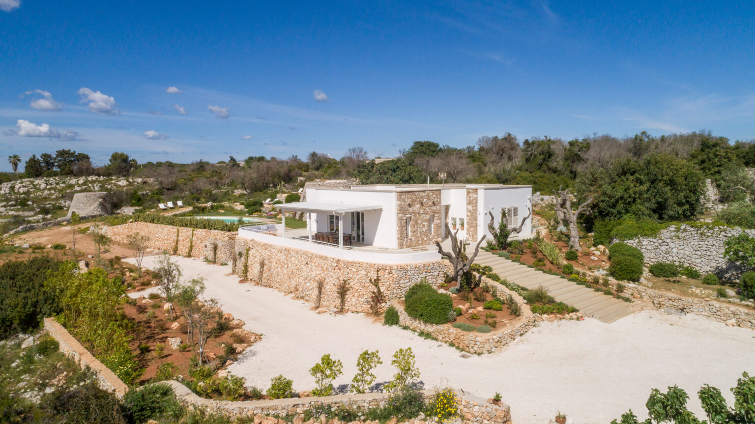 Modern villa surrounded by lush greenery and landscaped gardens, featuring a stone wall, outdoor seating area, and a swimming pool in a tranquil setting. Ideal for luxury retreats and vacation rentals. | Only on SopranoVillas