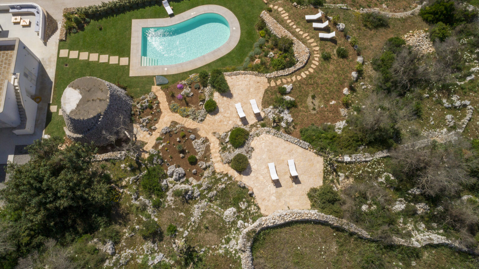Aerial view of a beautifully landscaped garden featuring a swimming pool, stone pathways, and lounge chairs, surrounded by lush greenery and rocky terrain. The image showcases a charming stone structure and well-maintained flower beds, highlighting a serene outdoor space perfect for relaxation. | Only on SopranoVillas