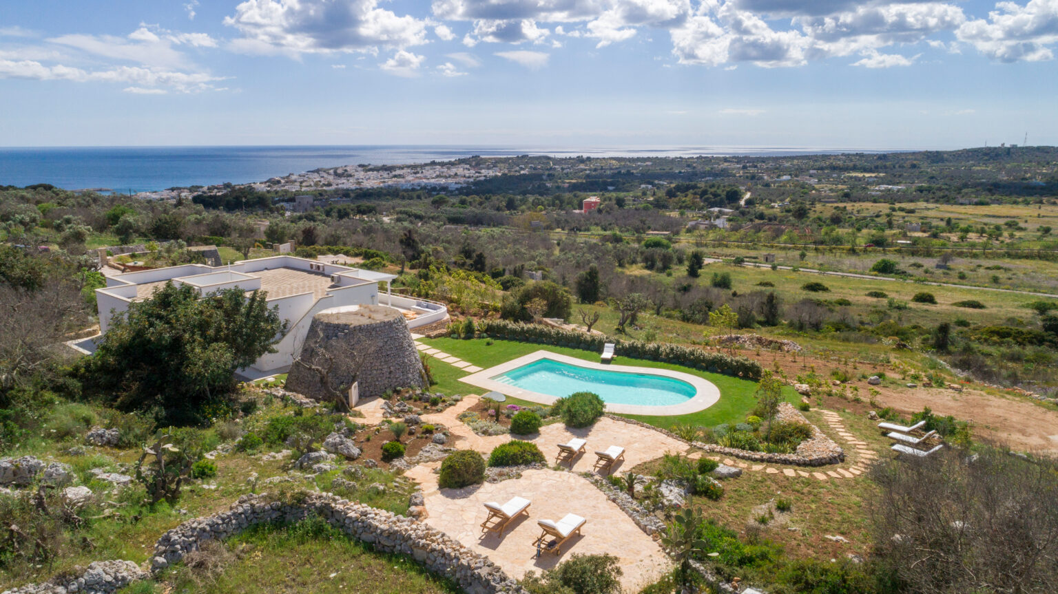 Aerial view of a luxurious villa with a swimming pool surrounded by lush greenery, overlooking the coastline and distant hills under a partly cloudy sky. The property features landscaped gardens and outdoor seating areas, ideal for relaxation and enjoying scenic views. | Only on SopranoVillas