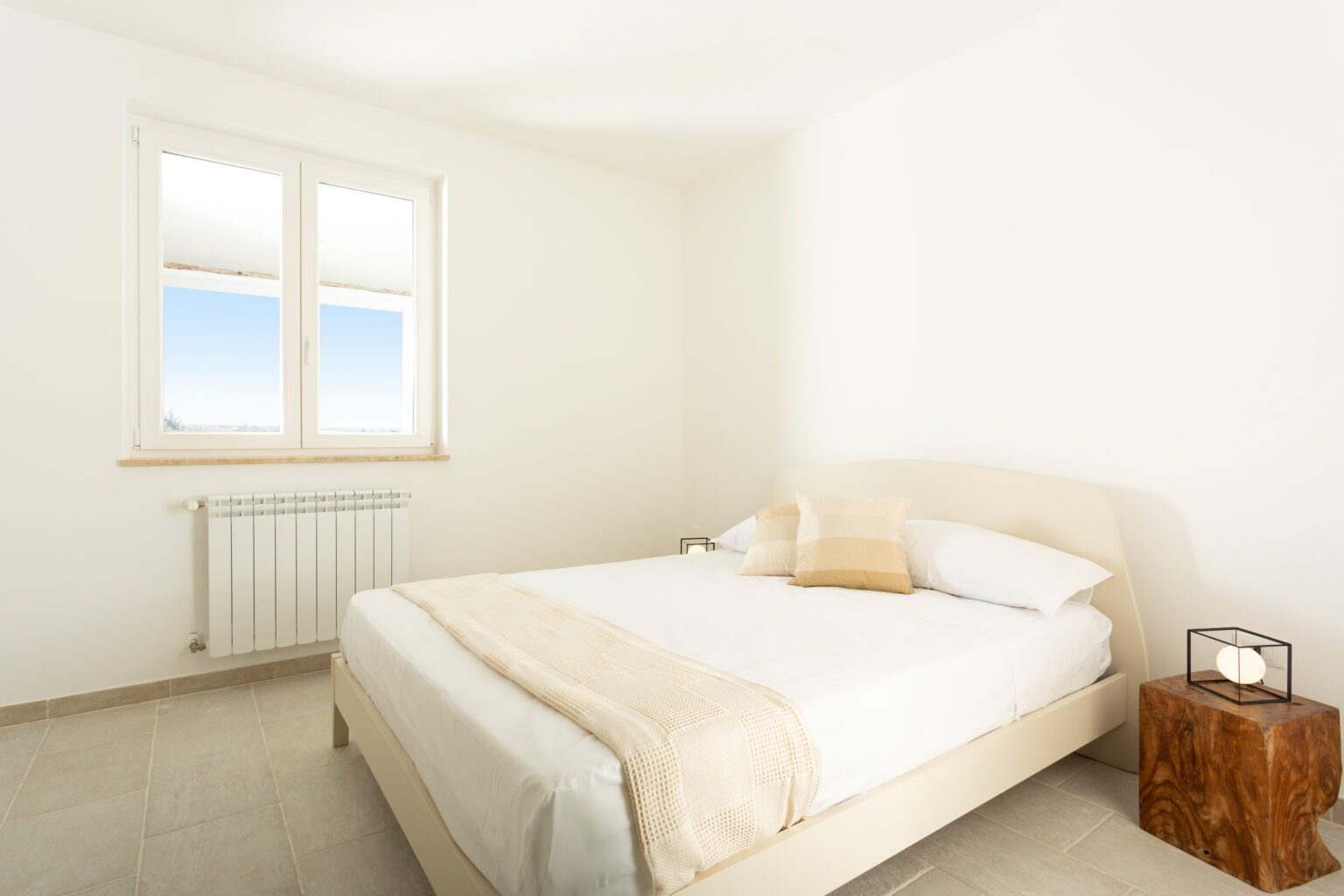 Bright and airy bedroom featuring a simple bed with a light-colored blanket, a wooden nightstand, and large windows allowing natural light to fill the space. | Only on SopranoVillas