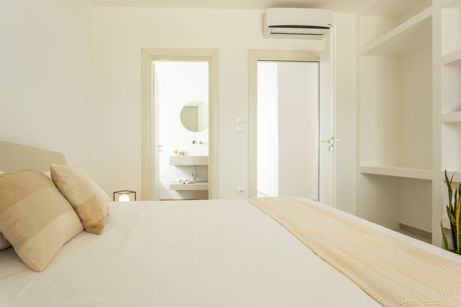 Cozy bedroom interior featuring a light color scheme, plush pillows on a neatly made bed, and a view of an adjoining bathroom through a door. The room includes built-in shelves and an air conditioning unit, enhancing comfort and functionality. | Only on SopranoVillas