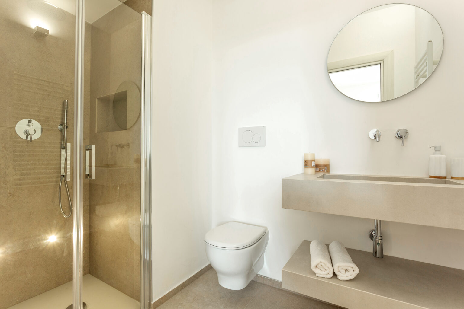 Modern bathroom featuring a walk-in shower, minimalist sink with a round mirror, and a toilet, complemented by neutral tones and soft lighting. | Only on SopranoVillas