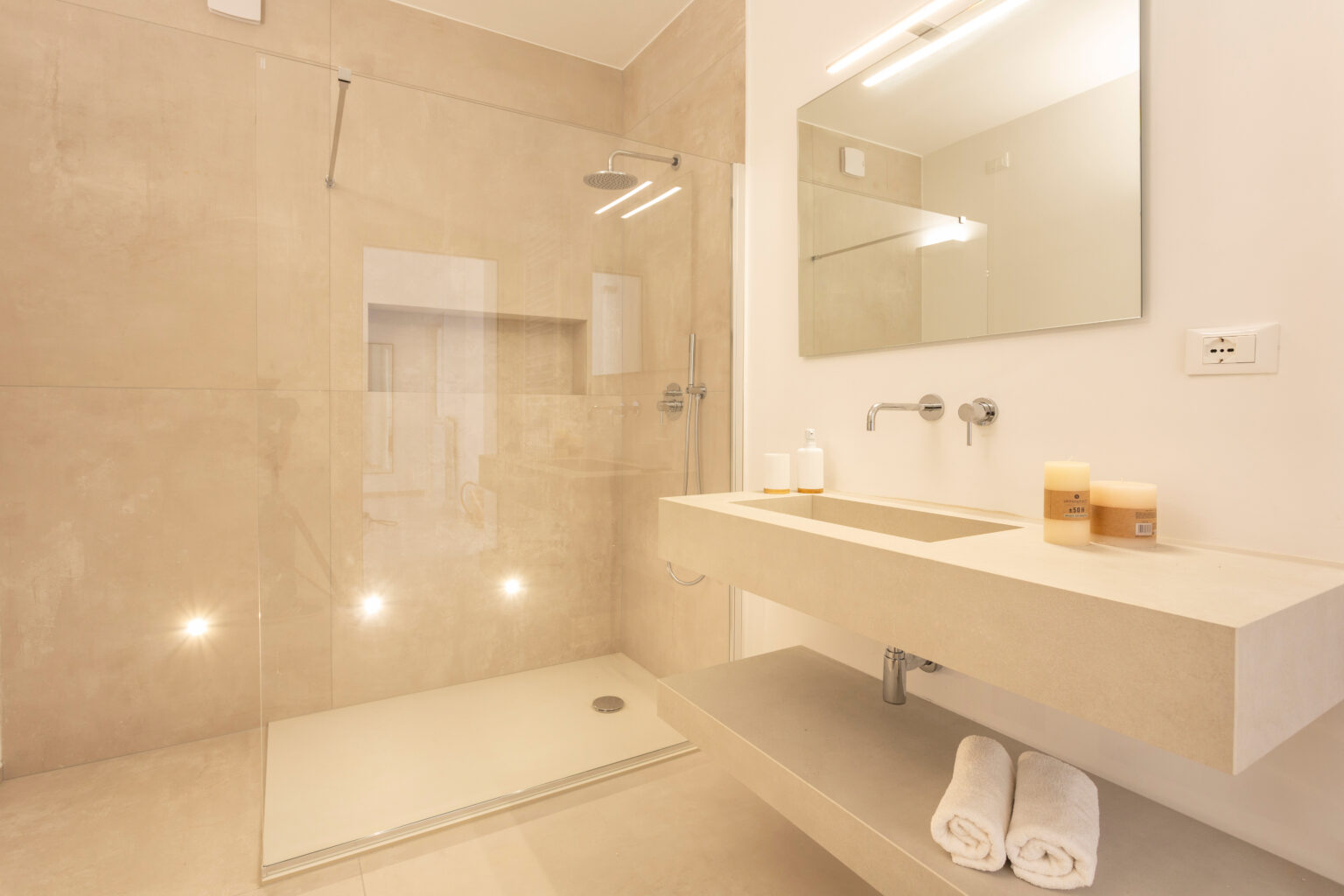 Modern bathroom featuring a walk-in shower with glass enclosure, minimalist sink with countertop, illuminated mirror, and neatly arranged towels and candles for a clean, contemporary aesthetic. | Only on SopranoVillas