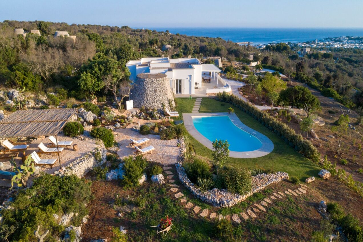 Aerial view of a modern villa with a swimming pool, surrounded by lush greenery and rocky terrain, overlooking the coastline. The property features outdoor lounging areas, landscaped gardens, and a natural stone structure, ideal for relaxation and scenic views. | Only on SopranoVillas