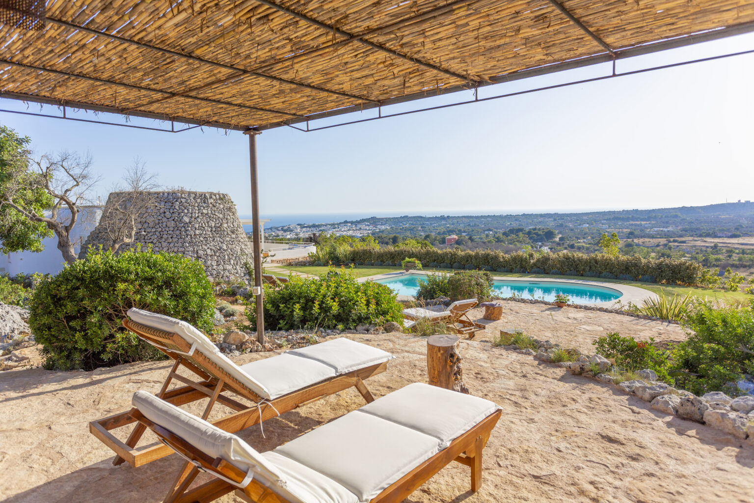 Scenic view of a tranquil outdoor space featuring wooden lounge chairs under a bamboo canopy, overlooking a swimming pool and lush greenery with a distant coastal landscape. Perfect for relaxation and leisure in a serene setting. | Only on SopranoVillas