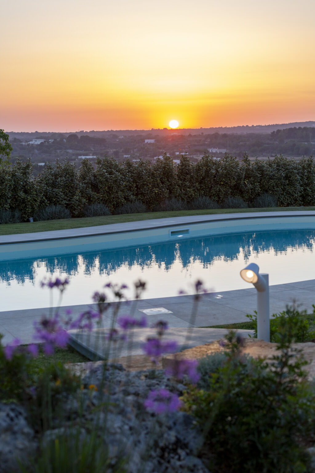 Serene outdoor swimming pool reflecting the sunset, surrounded by lush greenery and blooming flowers, creating a tranquil atmosphere. | Only on SopranoVillas