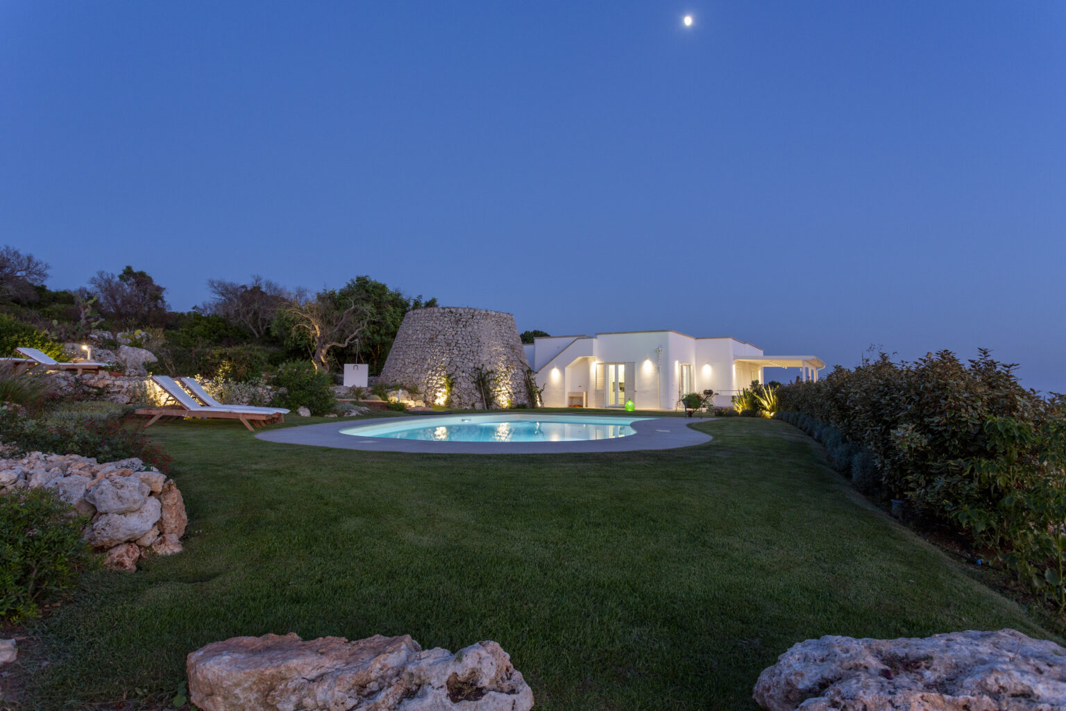 Modern villa with a swimming pool surrounded by lush greenery, illuminated at dusk, showcasing a serene outdoor living space. | Only on SopranoVillas