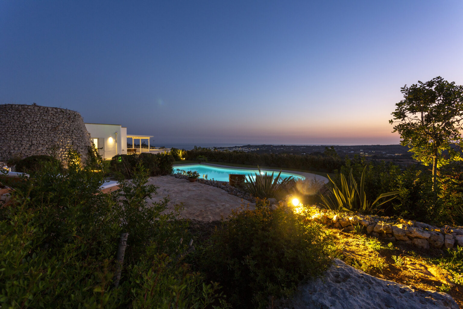 Scenic view of a luxury villa at dusk, featuring a glowing swimming pool surrounded by lush greenery and illuminated pathways, overlooking a tranquil landscape. | Only on SopranoVillas