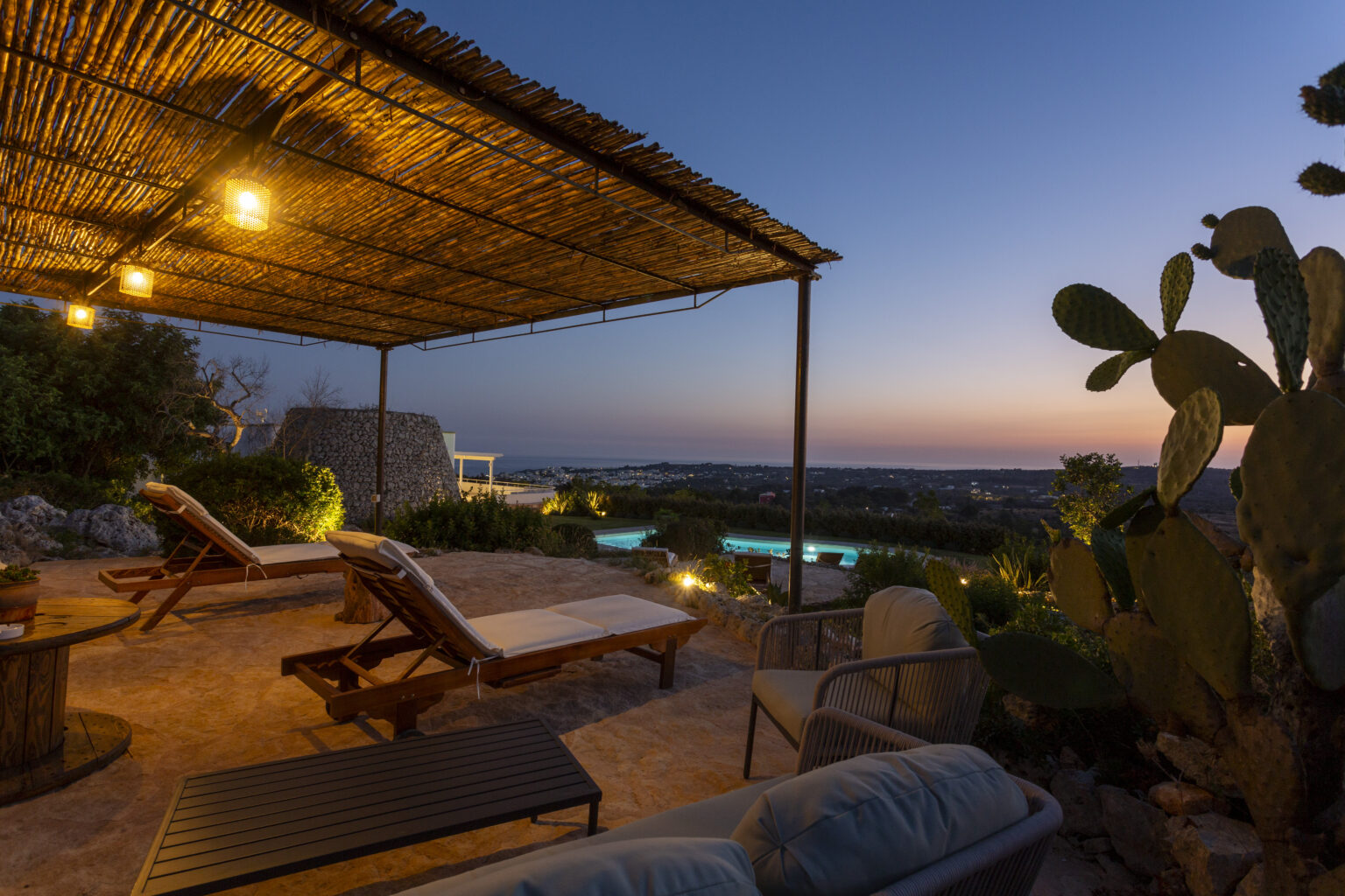 Cozy outdoor seating area with lounge chairs under a pergola, illuminated by warm lights, overlooking a serene landscape at sunset. | Only on SopranoVillas