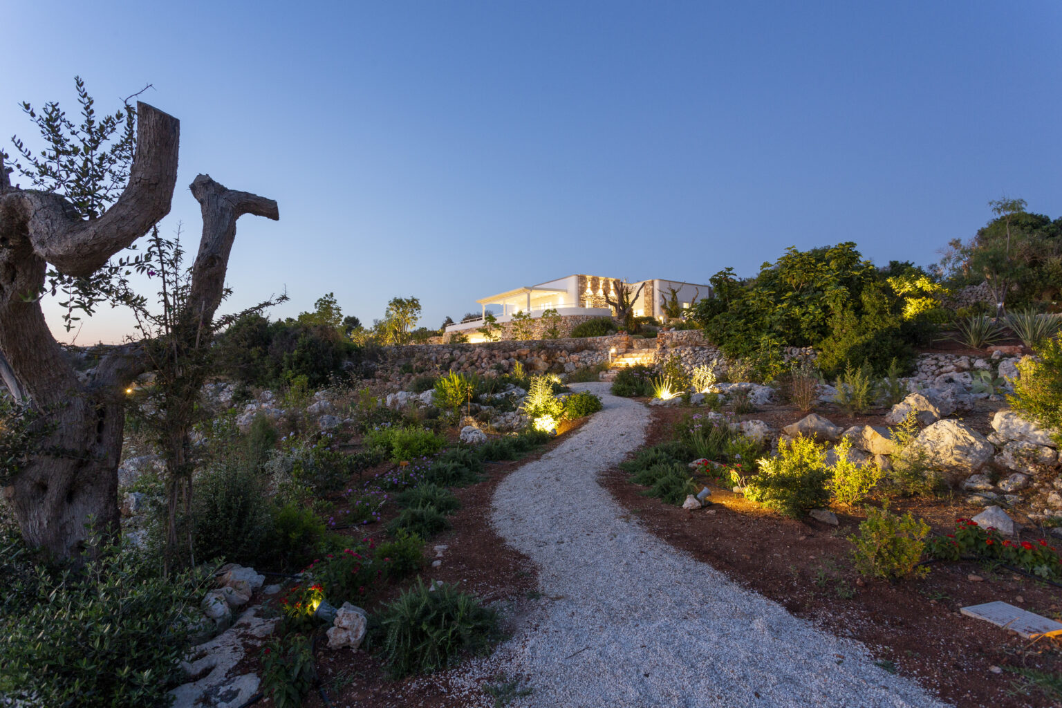 Scenic garden pathway winding through lush vegetation, illuminated by soft lights, leading to a beautiful building in the background under a twilight sky. | Only on SopranoVillas