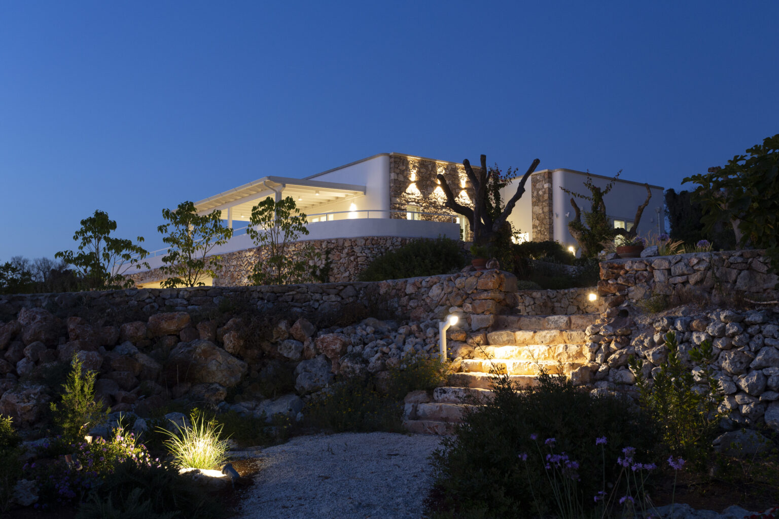 Modern architectural home illuminated at night, surrounded by landscaped stone gardens and decorative lighting. | Only on SopranoVillas