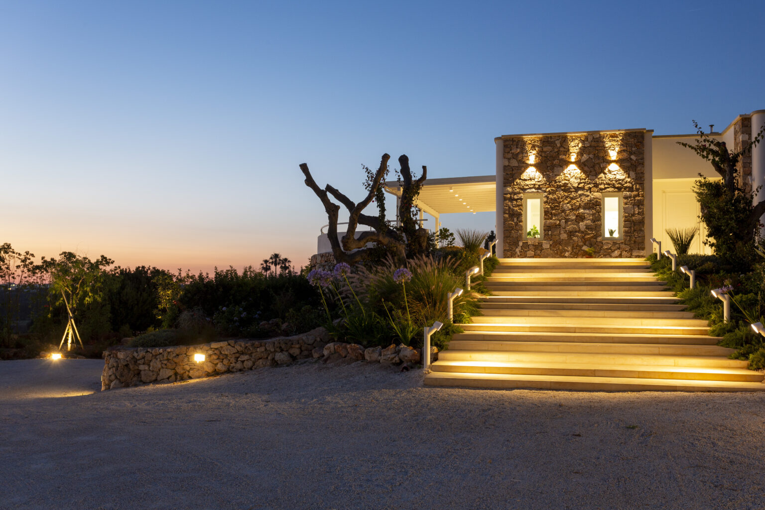 Modern architectural design of a stone entrance with illuminated steps leading to a stylish building at sunset, surrounded by landscaped greenery and trees. | Only on SopranoVillas