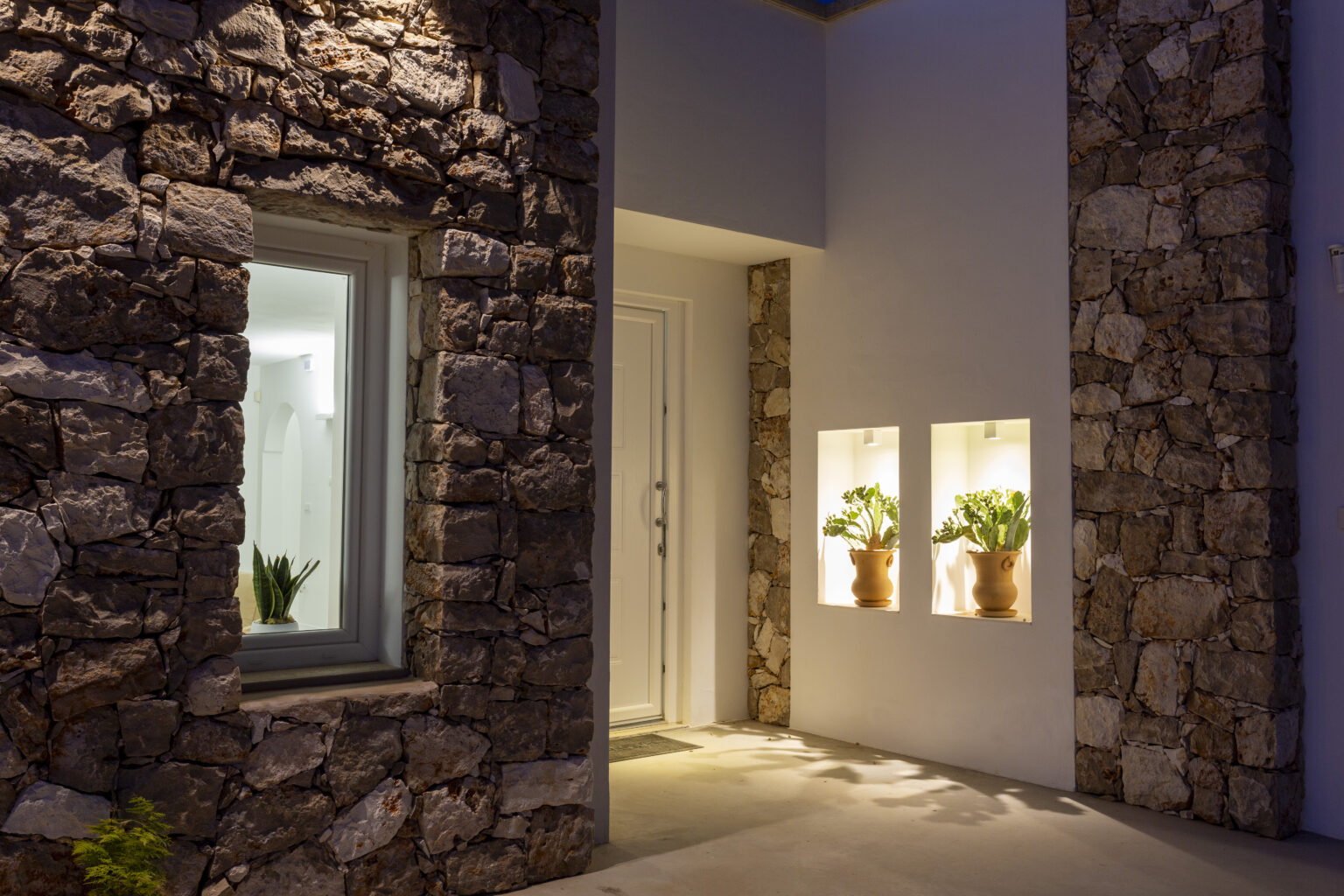 Modern entrance of a home featuring natural stone walls, illuminated potted plants in wall niches, and a sleek white door, creating a warm and inviting ambiance at night. | Only on SopranoVillas
