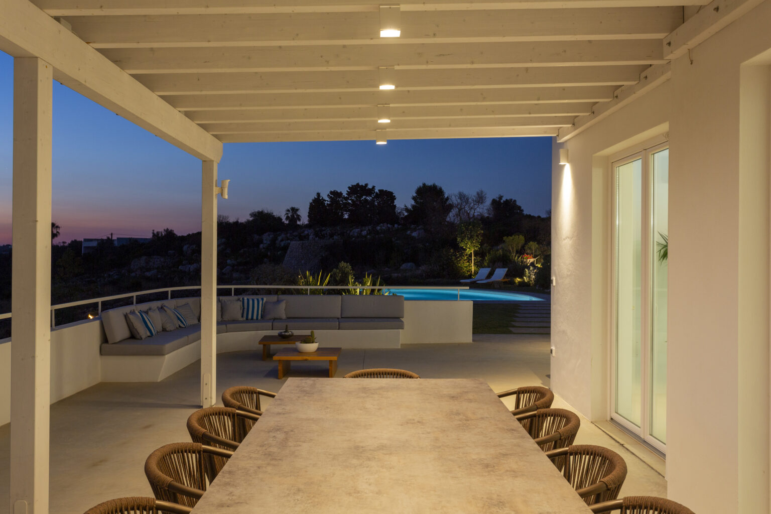 Modern outdoor dining area with a large stone table and comfortable chairs, illuminated by soft lighting, overlooking a serene pool and landscaped garden at twilight. | Only on SopranoVillas