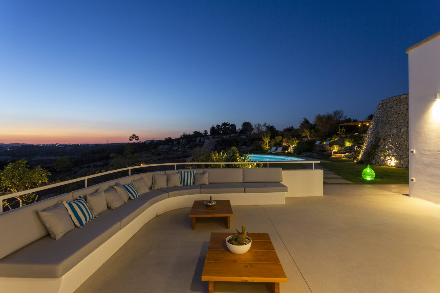 Modern outdoor terrace at dusk featuring a comfortable lounge area with cushions, a coffee table, and a serene view of the pool and landscape under a twilight sky. | Only on SopranoVillas