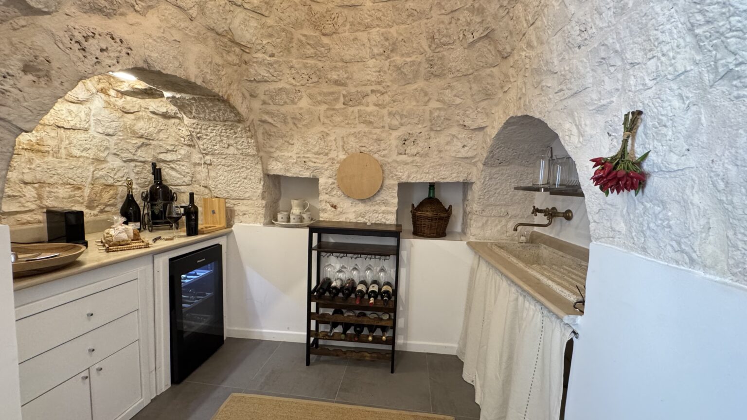 Cozy kitchen interior featuring arched stone walls, modern cabinetry, a wine rack, and a small refrigerator, creating a blend of rustic charm and contemporary design. | Only on SopranoVillas