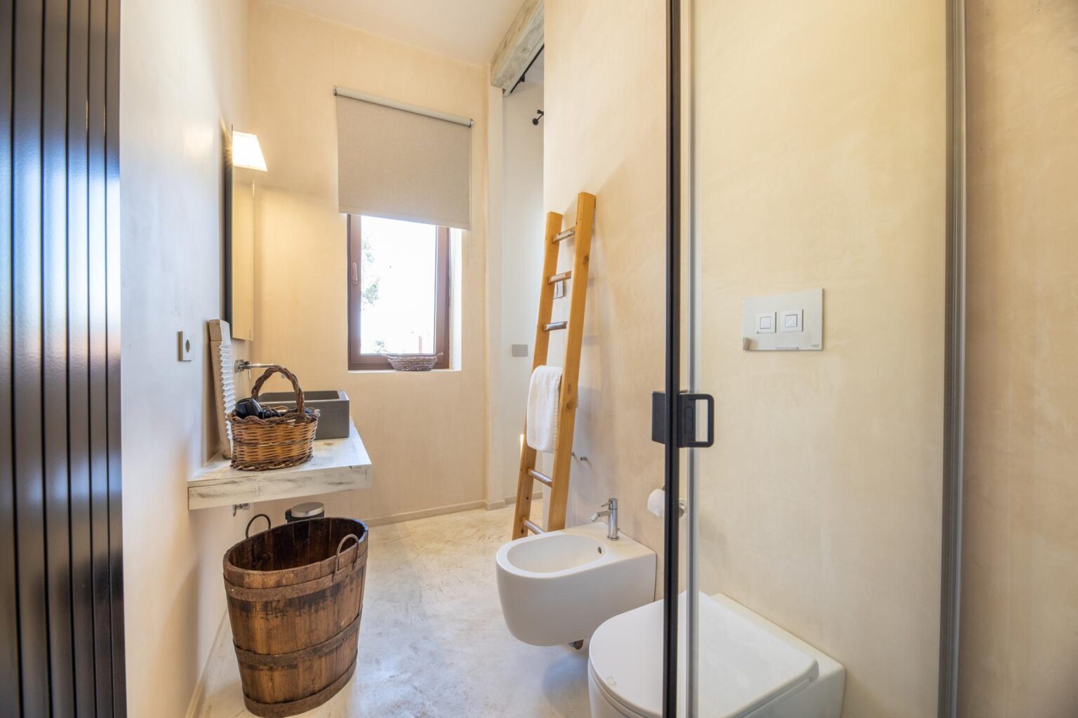 Modern bathroom with minimalist design featuring a glass shower, a wall-mounted toilet, a wooden ladder, and natural light from a window. Decorated with a wicker basket and a wooden trash bin, the space emphasizes simplicity and functionality. | Only on SopranoVillas