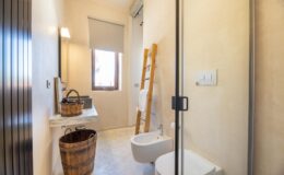 Modern bathroom with minimalist design featuring a glass shower, a wall-mounted toilet, a wooden ladder, and natural light from a window. Decorated with a wicker basket and a wooden trash bin, the space emphasizes simplicity and functionality.