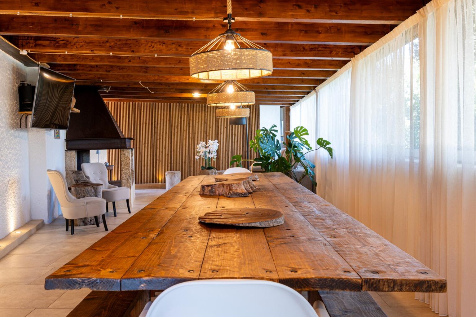 Modern dining room featuring a large wooden table, elegant pendant lights, and natural decor elements. The space is bright with large windows and plants, creating a cozy and inviting atmosphere. | Only on SopranoVillas