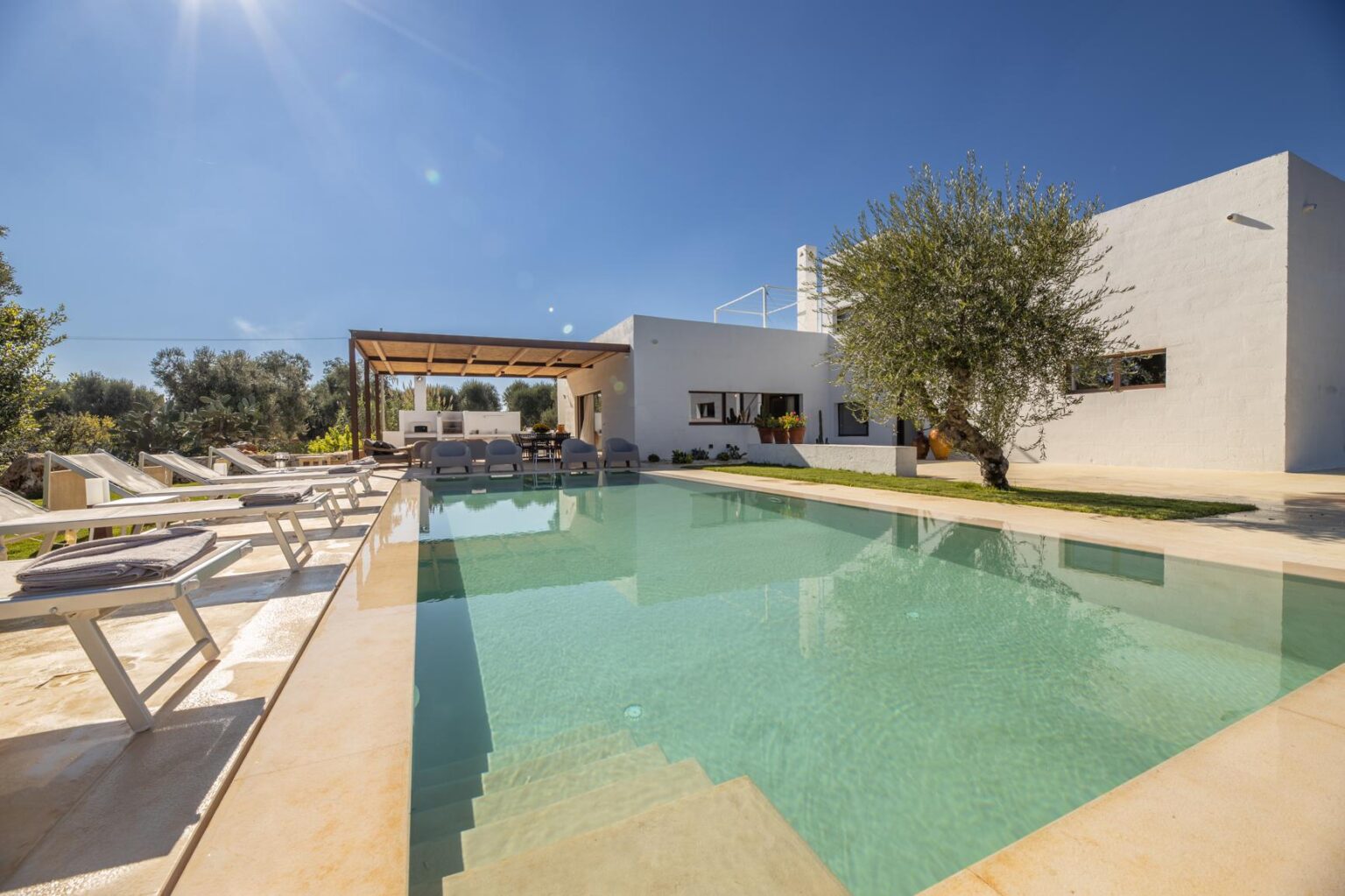 Modern villa with a swimming pool surrounded by lounge chairs and olive trees, set under a clear blue sky. Ideal for luxury vacation rentals or outdoor gatherings. | Only on SopranoVillas